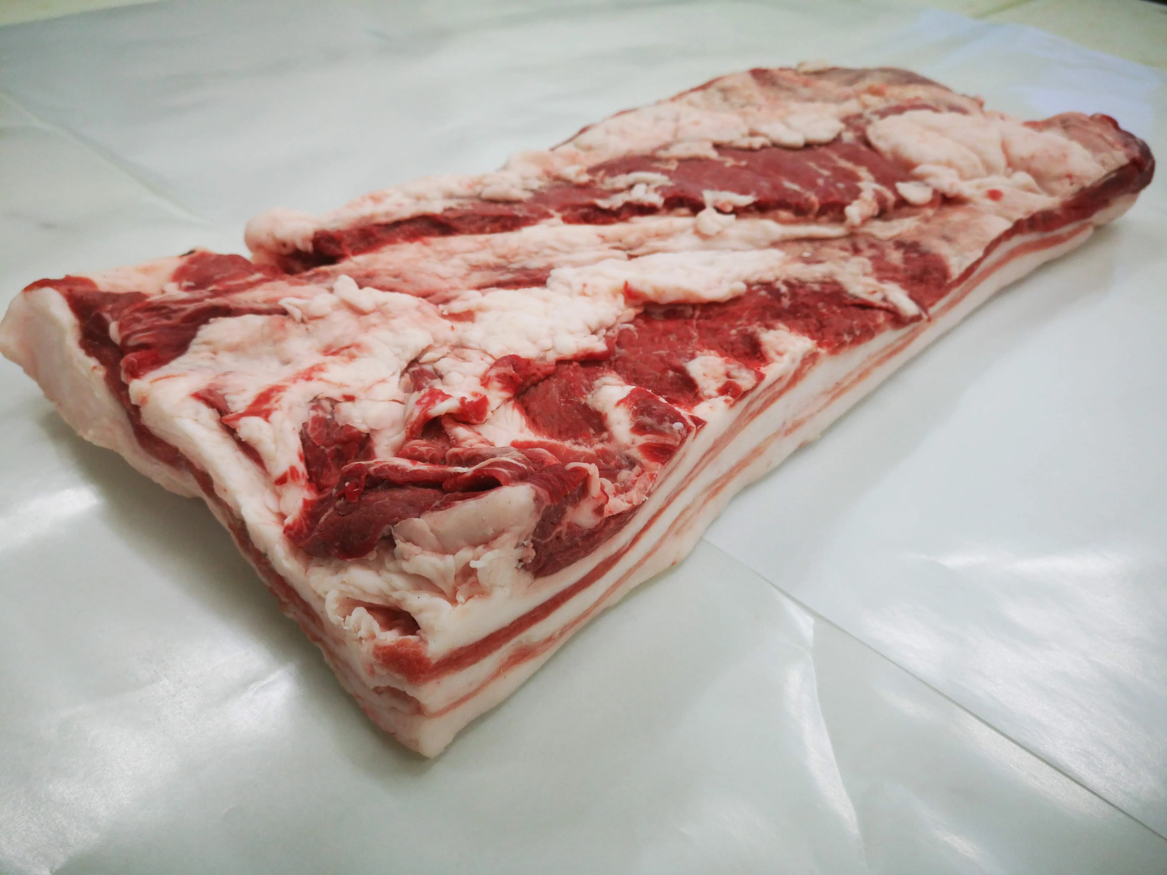 A single pork belly laid skin-down on white paper, showing the alternating layers of lean and fat along the cut edge.